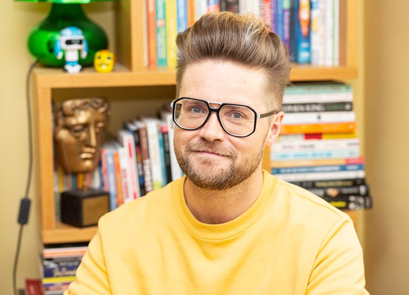 Myles McLeod in a yellow jumper, in front of a bookshelf with a BAFTA mask